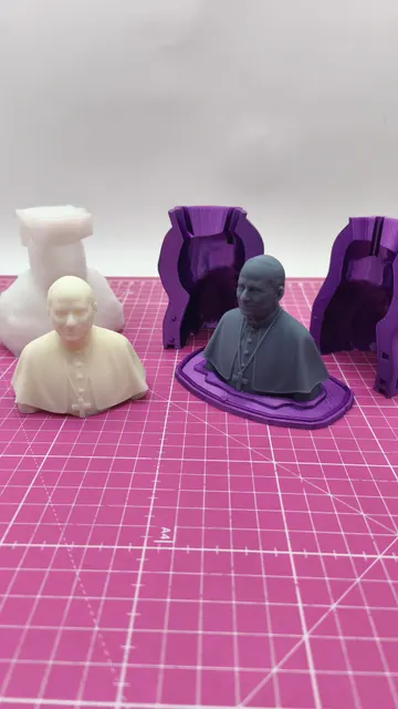 A cream and a grey printed pope bust standing in front of the purple two-part molds and the white silicone negative they came from. Made by @punto3dperu.