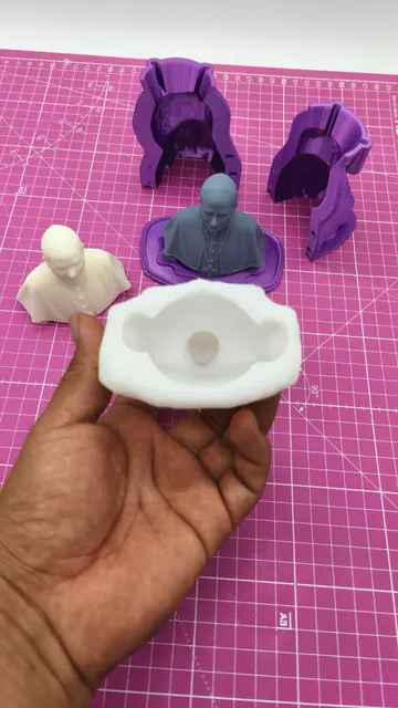 A white silicone negative of a pope bust held up in front of the purple printed molds and the finished casts. Made by @punto3dperu.