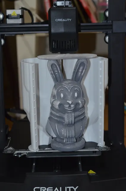 A grey 3D-printed rabbit bust with a knitted scarf, dry-brushed white to show the detail, standing in front of the open white two-part mold printed for it. Made by @zevra.home.