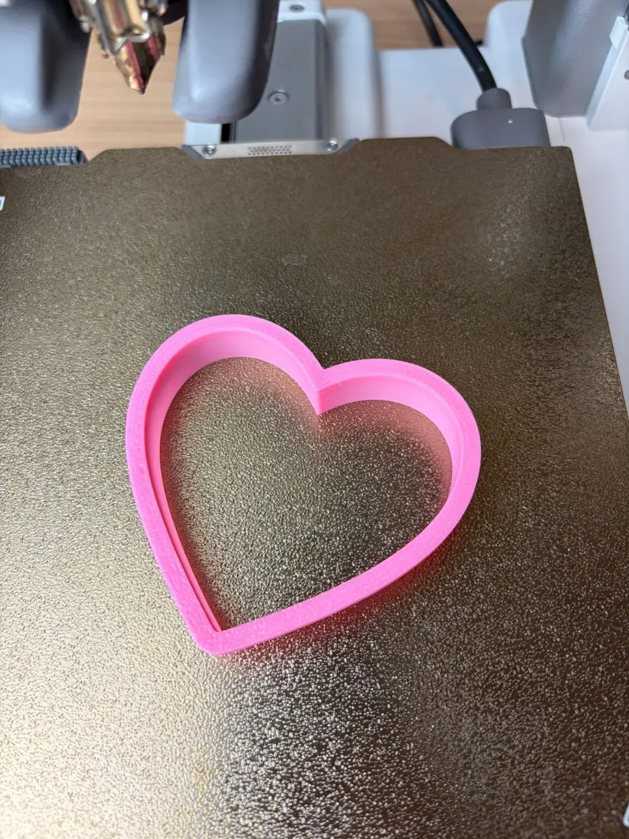 The pink 3D-printed heart cookie cutter on a textured print bed, just off the printer