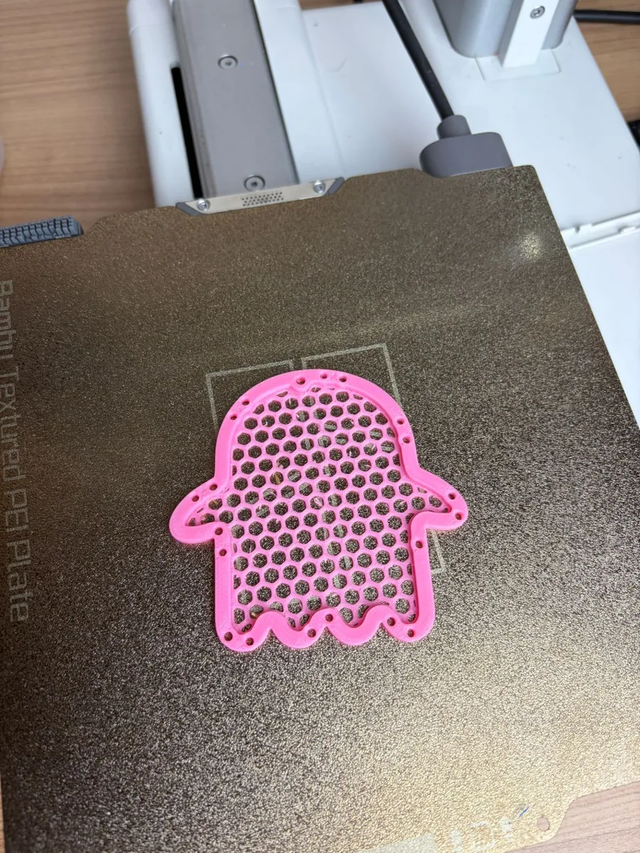 A pink honeycomb mesh ghost net fresh off the printer, lying flat on a textured print bed with the joint holes round its rim