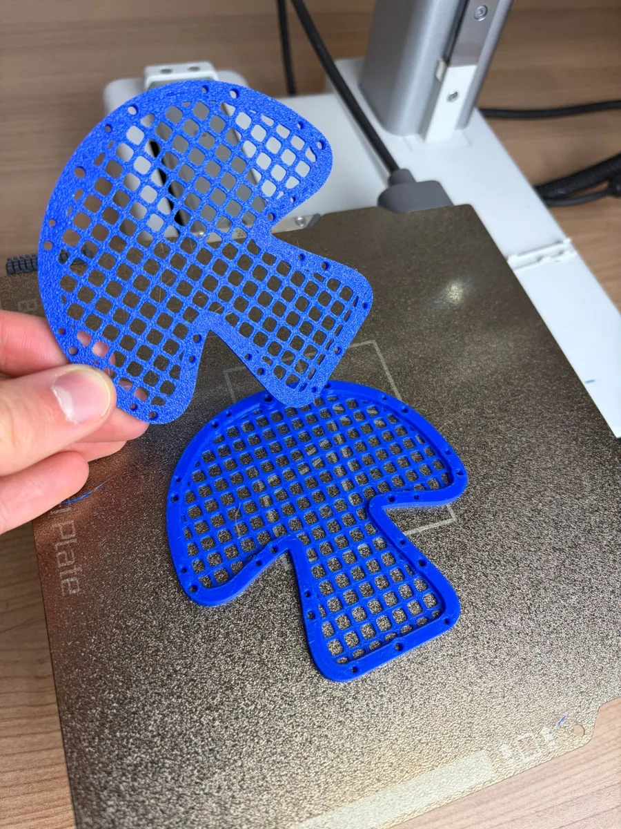 Two blue square-mesh mushroom nets fresh off the printer, one held up in a hand, the other lying on the print bed with the pin holes round its rim