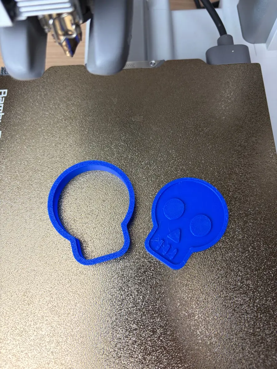 The blue 3D-printed skull emoji cookie cutter and its face stamp side by side on the print bed