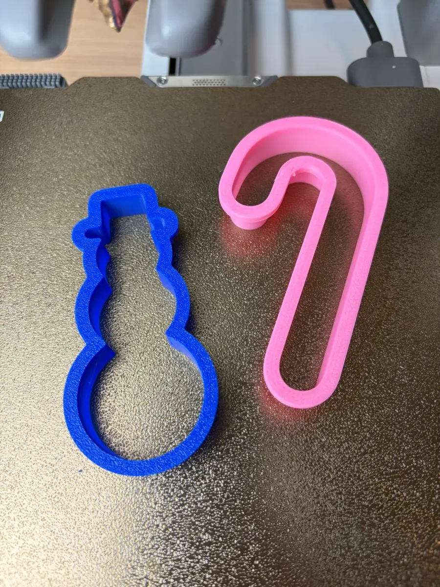 The blue snowman cookie cutter next to the pink candy cane cookie cutter, both 3D printed, on the print bed